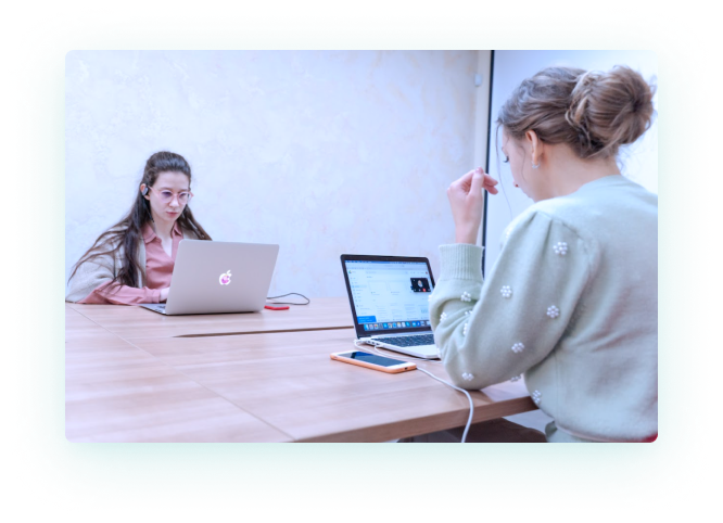 Two Girls Sitting Behind Light Wooden Table Looking into Their Laptops